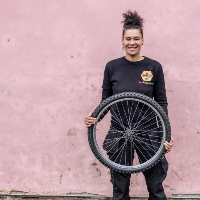 Greater Manchester's Bicycle Mayor, Belinda Everett, stood smiling against a pastel pink wall holding a bike wheel in one hand, wearing all black.