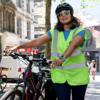 A woman in a high-vis and helmet smiles as she stands next to her bike.
