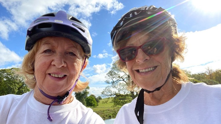 A close up selfie of two women smiling on a sunny day with blue skies in the background. The friends are both wearing white t-shirts. The photo was taken during their journey on the Bay Cycle Way, Lancaster.