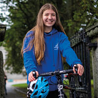 Molly, wearing a blue jacket, smiling as she stands with her bicycle