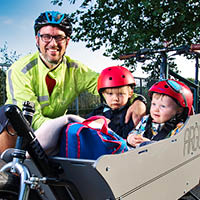 Richard smiling with his two children sitting in a cargo bike trailer