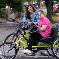 A mother on her cycle and a young adult who is her daughter on an adapted cycle both smiling on a residential road in Liverpool City Region 