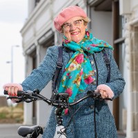 An older woman dressed in bright colours smiling and stood holding on to the handlebars of her cycle 