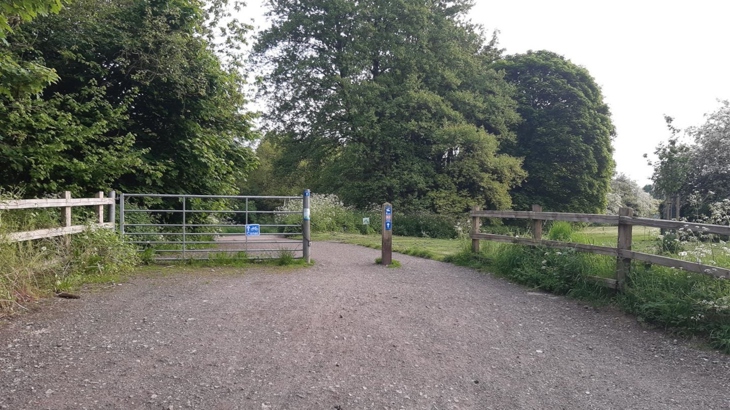 The wide entrance to a traffic-free path has a metal gate across half of it, the other half is open with a wooden post at the centre of the gap. National Cycle Network signs are on both the gate and the post. The path beyond is tree lined, some larger tress are in the distance and green shrubs are in the foreground. The scene is lush and green with plants in full leaf.
