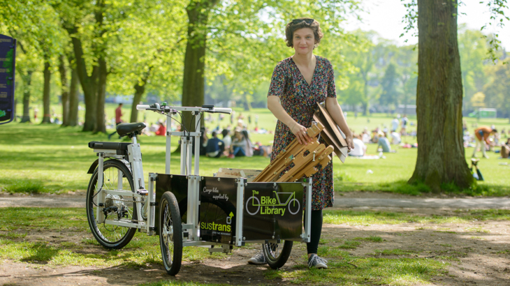 A woman is unloading some wooden equipment from the front of a cargo bike. The scene is of a very busy park on a warm sunny day. There are trees just behind her and beyond is a wide open space where hundreds of people are relaxing.