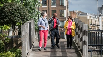 Three people are in conversation as they walk over a small wooden bridge in Tower Hamlets, London. A leafy green tree is to the left of them and tall buildings are in the distance.