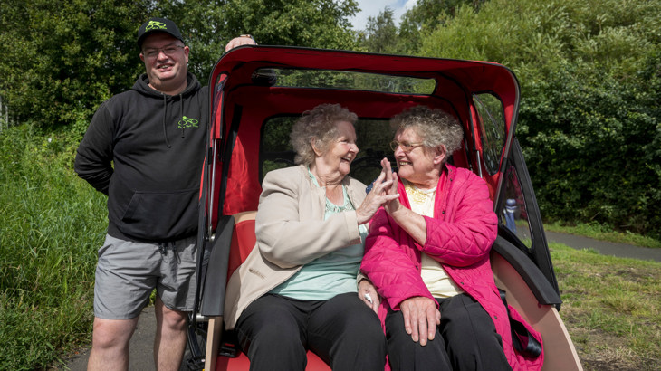 Two members of the Forever Young group pictured smiling and touching hands as they sit aboard a trishaw