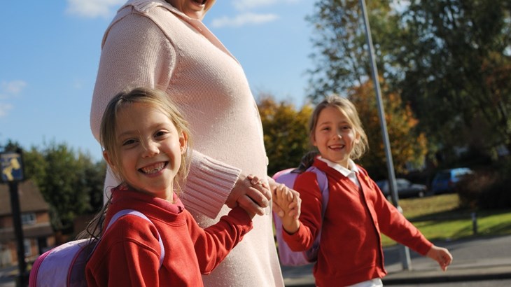 Mum walking to school, holding hands with her twin daughters smiling at the camera