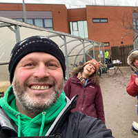 Sustrans Walking and Cycling Technical Lead, Wayne Brewin, smiling and taking a selfie-style photo with two colleagues in front of bicycle storage in the school playground.