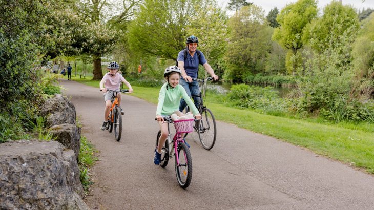 Two children and a man cycle on a traffic-free path surrounded by greenery.