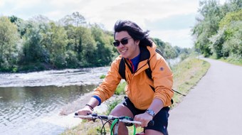 Cyclist in dark glasses and orange jacket on asphalt cycle path by river