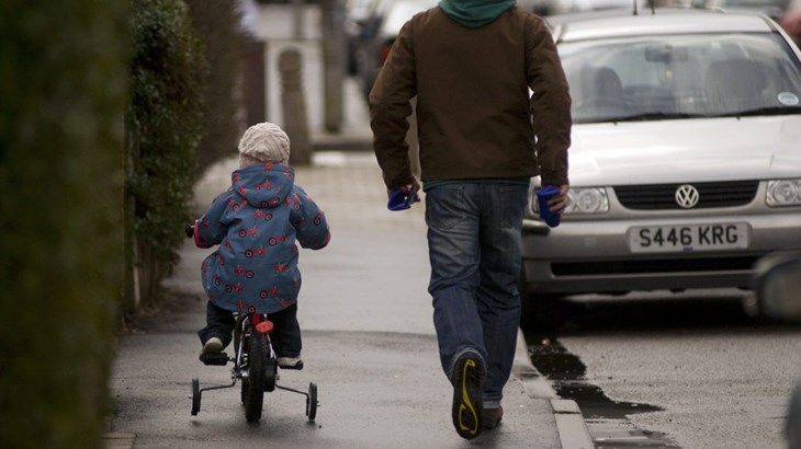 A Man Walking On Pavement While Child Cycles