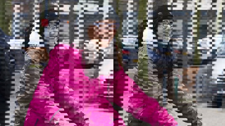 A female cycling in a pink coat and woolly hat.