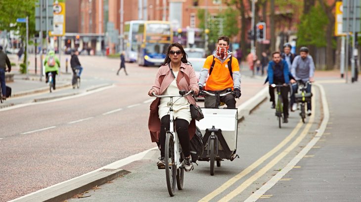 Woman wearing a pink jacket and sunglasses cycling down a segregated cycle lane in Manchester city centre with a man on a cargo bike behind her.