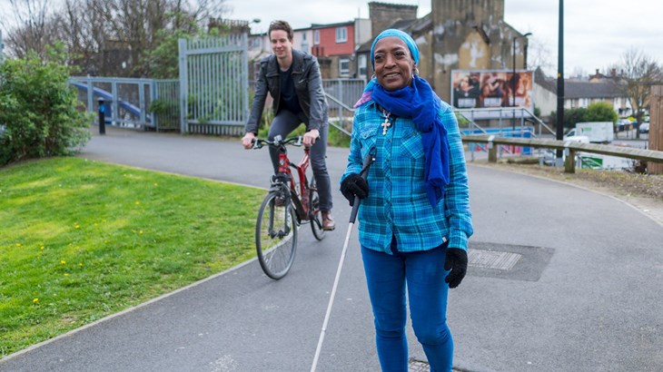 Lady in bright blue using a cane walking along a traffic-free path with a male cycling behind her.