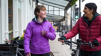Two women wearing brightly coloured raincoats, chatting and walking with their bikes through a high street