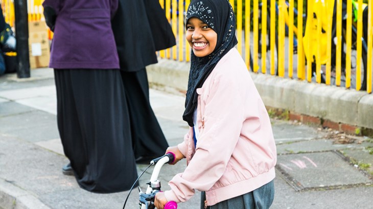 Secondary school girl in a headscarf standing with her bicycle outside the school gate.
