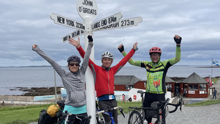 A group of three adults dressed in cycling gear with their bikes stood with their hands in the air looking victorious next to the John'o Groats signpost by the coast in Scotland